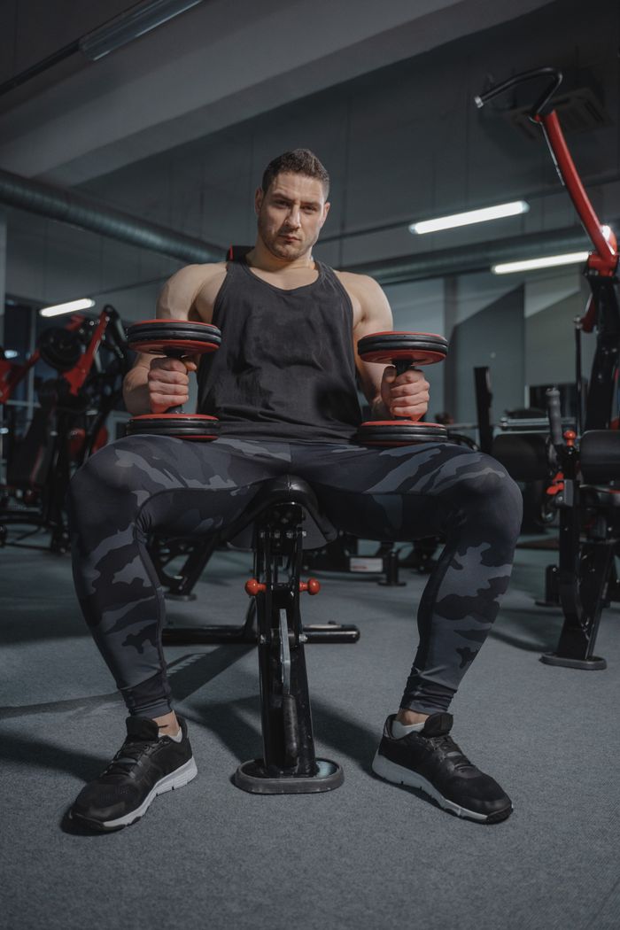Muscular man wearing activewear lifts dumbbells while sitting in a gym, showing strength and fitness.